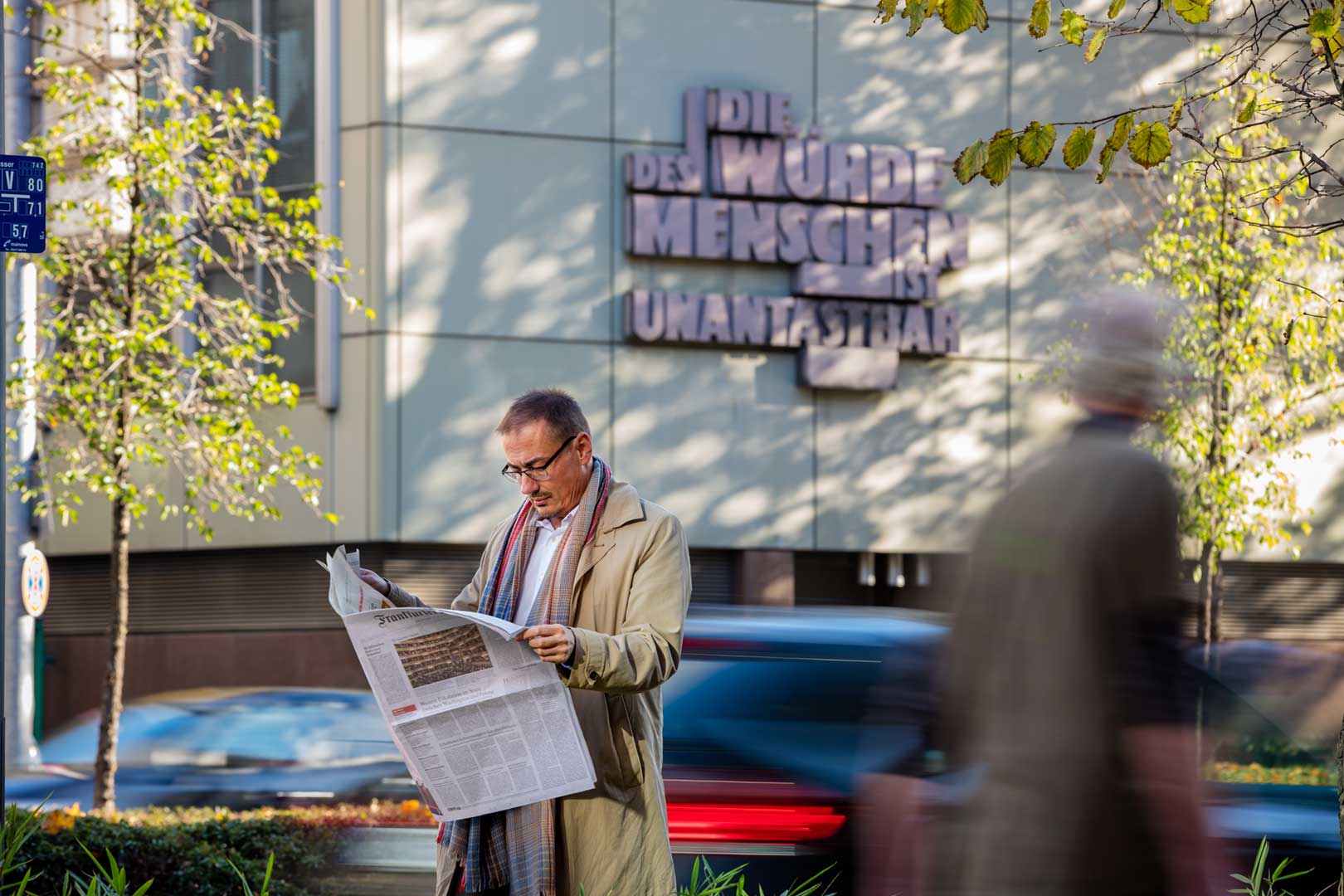 Mann im Mantel liest Zeitung auf der Straße; unscharfe Fußgänger und Gebäude mit deutschem Schild im Hintergrund.